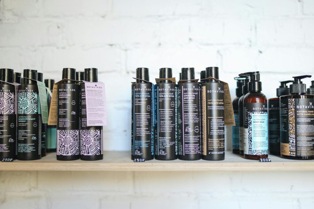 A variety of natural cosmetic bottles lined up on a wooden shelf against a white brick wall, suggesting eco-friendly beauty.